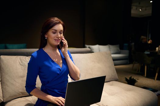 Confident European business woman talking on mobile phone while working on laptop in an interior design studio. Professional designer giving online consultation to clients about a new housing project