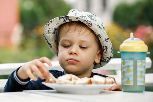 The boy eats toast for breakfast with juice in the background of a summer garden. A plastic cup with pineapple juice on the table.