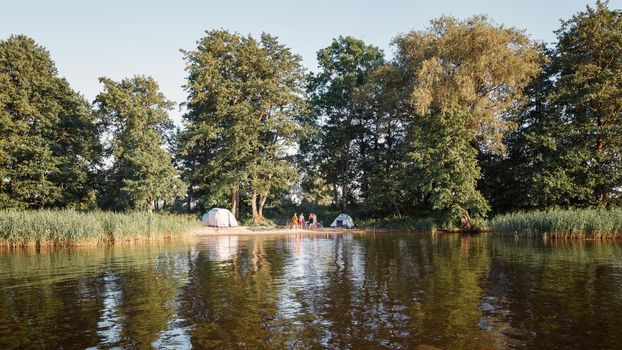 Summer camping on lake shore. Group of four young tourists standing near tent under beautiful green trees. Tourism, friendship and beauty of nature concept