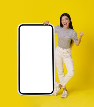 Full body asian girl stand near huge, giant smartphone with white screen happy making yeah, winner hand gesture dressed in casual isolated on yellow background. Free space mock up.