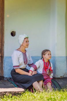 mother and daughter in Ukrainian national costumes are sitting near an old house