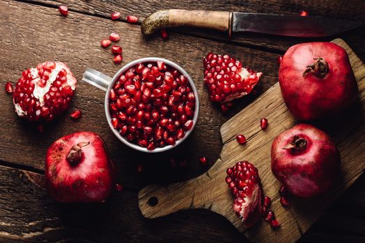 Metal mug full of pomegranate seeds. Whole fruits and pomegranate pieces on rustic wooden table. View from above