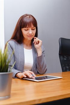 A brunette businesswoman in a gray jacket at her desk with a tablet in her hands. Business portrait in the office.