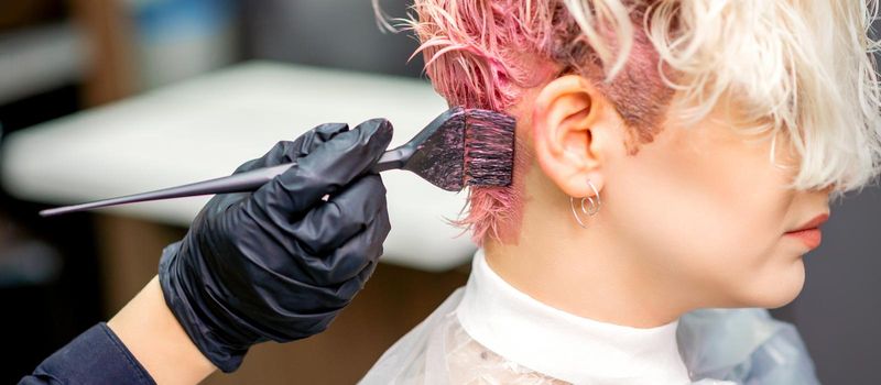 Hand of hairdresser dyeing hair in pink color by paintbrush on back of head of young caucasian woman in beauty salon. Selective focus