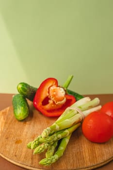 Vegetables lie on a wooden board: tomatoes, asparagus, cucumbers, red bell peppers. brown, light green background. place for text