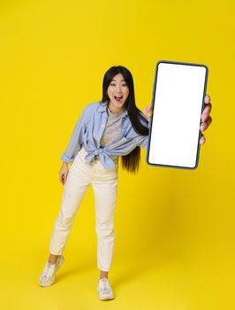 Young asian girl in casual happy holding smartphone showing white screen mobile app advertisement and excited smile on camera isolated on yellow background. Great offer. Product placement.