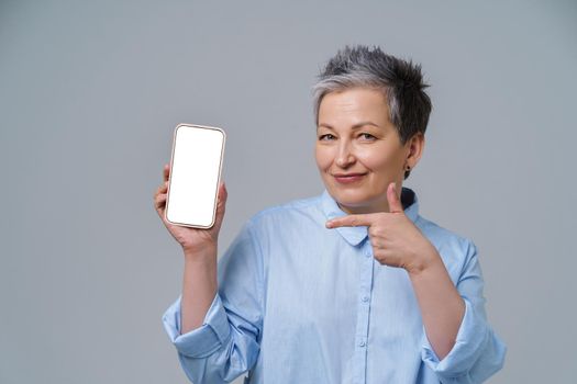 Mature grey hair woman smiling holding smartphone pointing on white screen working or shopping online, social media. Pretty woman in blue shirt isolated on white background.