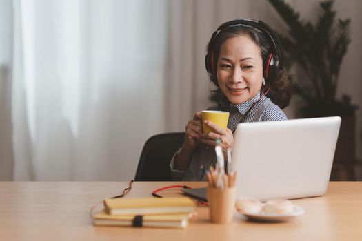 Portrait of Asian Senior Business woman wear headphone and with coffee using video call at morning at home. Account and Finance concept