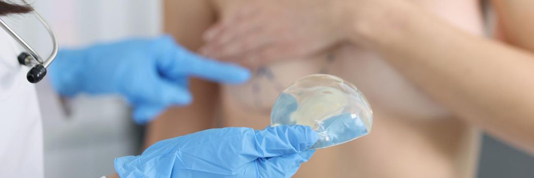 Close-up of doctor hold silicone breast implant and show marking on clients body. Woman sit topless in cabinet. Surgery, beauty, medicine, plastic concept