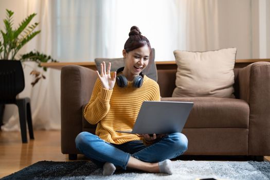 Happy young asian teen girl sit on sofa at home holding laptop looking at screen waving hand video calling
