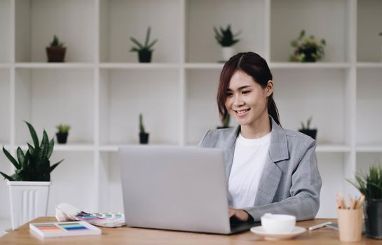 Cheerful attractive young female graphic designer smiling and working on laptop computer at her desk in modern office.