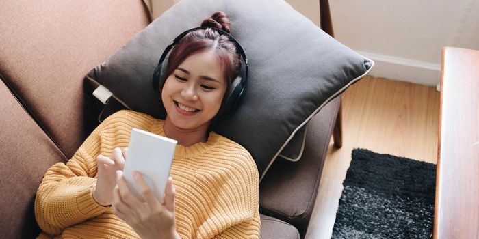 Lazy young woman spend weekend at home lying down on couch wearing headphones looking at smart phone screen listening music online.
