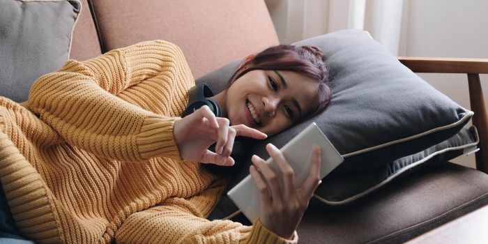 Lazy young woman spend weekend at home lying down on couch wearing headphones looking at smart phone screen listening music online.