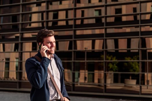 smiling young businessman walking by an office building in the financial district talking on the phone, concept of entrepreneurship and business, copy space for text