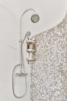 Shower faucet hanging on a white tiled wall and a shelf with toiletries in a bright washroom in a modern apartment