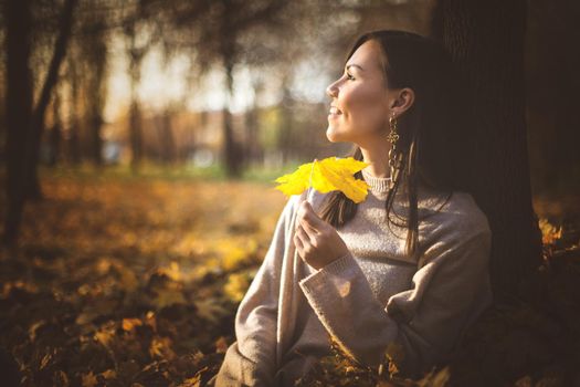 Young happy mixed race woman sitting near tree in autumn park at sunset.