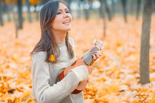 Young beautiful mixed race woman playing ukulele and singing in autumn park.