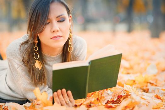 Central asian woman lying on fall foliage and reading book in park.