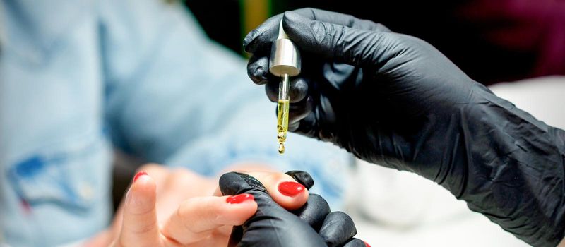 Manicure master applying oil with pipette to cuticle of female nails in nail salon