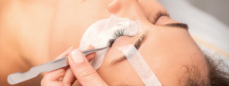 Young caucasian woman having eyelash extension procedure in beauty salon. Beautician glues eyelashes with tweezers