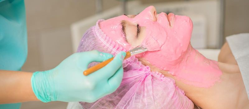 Hand of cosmetologist applying pink alginic mask to face of young woman in beauty salon