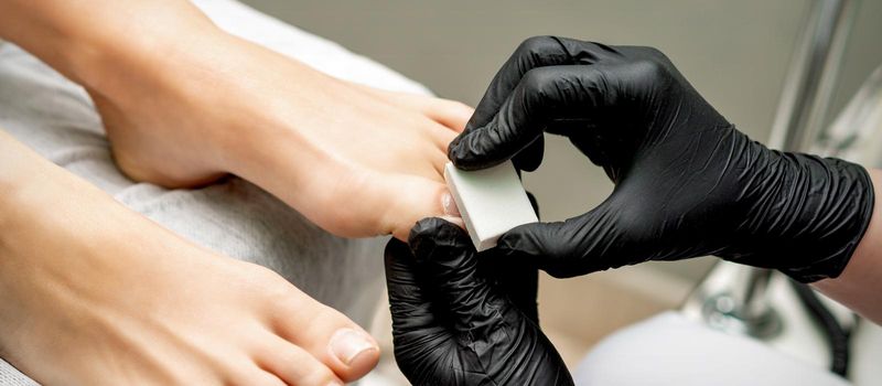 Hands of pedicurist buffing toenail of woman by white nail buff in nail salon