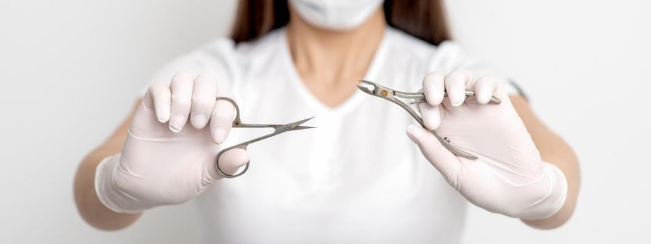 Manicure tools in hands of female manicurist wearing white mask and gloves on white background