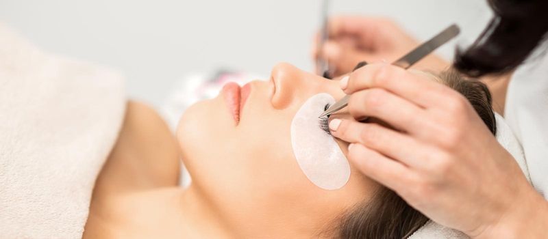 Young caucasian woman having eyelash extension procedure in beauty salon. Beautician glues eyelashes with tweezers