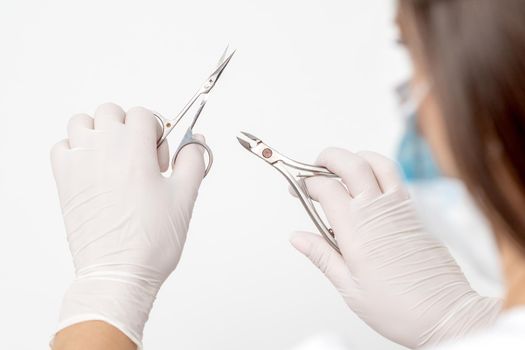Manicure tools in hands of female manicurist wearing white mask and gloves on white background