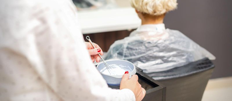 Hairdresser is preparing white dye in container on female client background