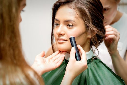 Makeup artist applying cream blush stick foundation on cheek of young caucasian woman
