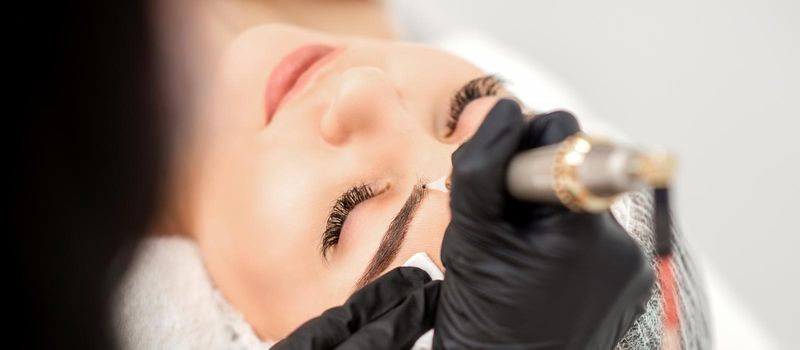 Beautician applying permanent makeup on eyebrows of young woman by special tattoo machine tool