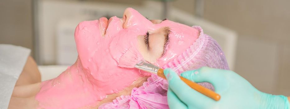 Hand of cosmetologist applying pink alginic mask to face of young woman in beauty salon