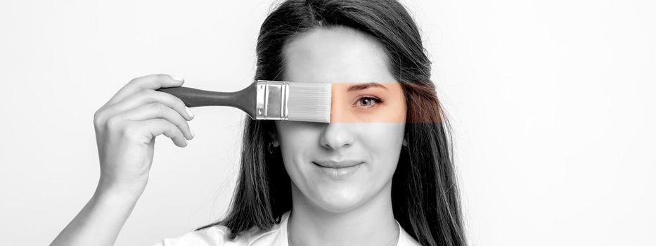 Black and white portrait of young woman painting her eye in yellow color with paintbrush on white background