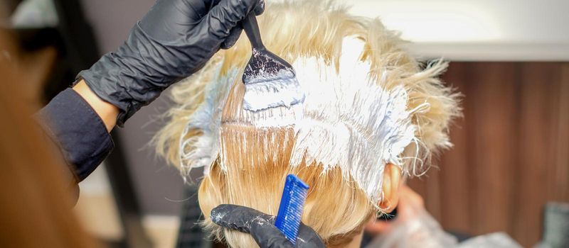 Back view of hands of hair stylist dyeing hair of young woman in white color in hair salon