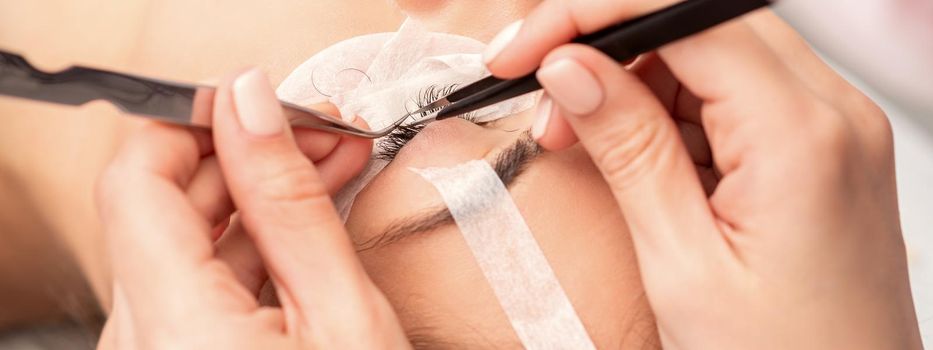 Young caucasian woman having eyelash extension procedure in beauty salon. Beautician glues eyelashes with tweezers