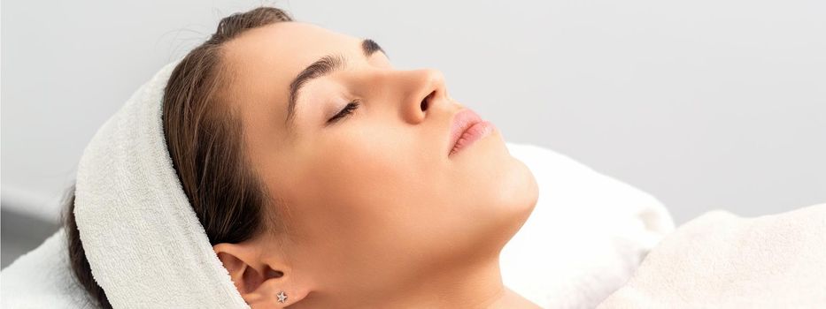 Side view of young woman lying on beautician table with closed eyes while waiting for cosmetic procedure in beauty salon