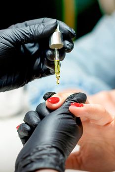 Manicure master applying oil with pipette to cuticle of female nails in nail salon