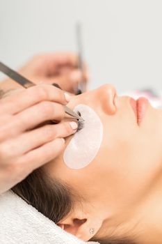Closeup view of young caucasian woman having eyelash extension procedure in beauty salon. Beautician glues eyelashes with tweezers