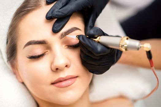 Beautician applying permanent makeup on eyebrows of young woman by special tattoo machine tool