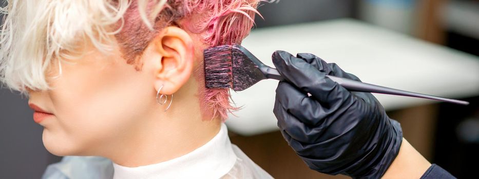 Hand of hairdresser dyeing hair in pink color by paintbrush on back of head of young caucasian woman in beauty salon. Selective focus