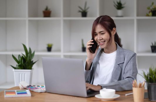 Asian woman designer working using a smartphone blank white computer screen placed at table with color samples