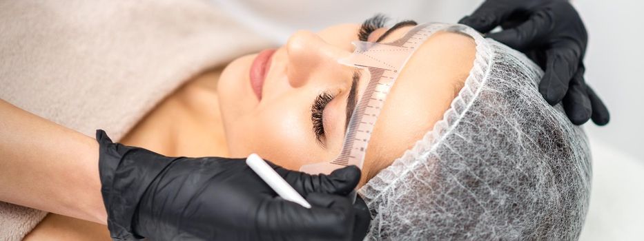 Cosmetologist is measuring with ruler the brows of young caucasian woman before permanent makeup tattoo