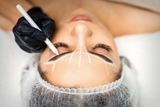 The beautician marks the eyebrow with a white pencil to prepare a permanent makeup procedure
