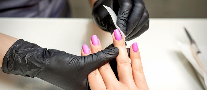 Manicurist is cleaning pink nail polish on female fingernails after manicure procedure in nail salon