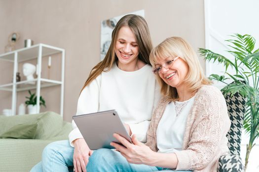 Senior woman and daughter playing with tablet pc. Smiling older woman and her granddaughter sitting home using gadget digital device for online shopping internet surfing video conference calling.