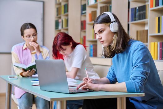 Group of teenage students study sitting at desk in library, in focus is guy in headphones using laptop book and writing in notebook. College, high school, education, learning, knowledge concept