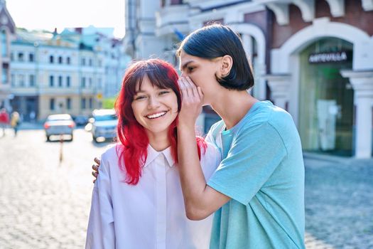 Couple of teenage students friends, guy whispers secret in ear of girlfriend on summer city street. Adolescence, friendship, relationships, surprise, emotions, youth concept