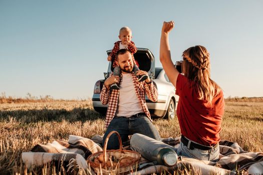 Happy Young Family Mom and Dad with Their Little Son Enjoying Summer Weekend Picnic Sitting on the Plaid Near the Car Outside the City in Field at Sunny Day Sunset, Vacation and Road Trip Concept