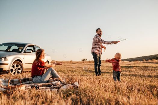 Happy Young Family Mom and Dad with Their Little Son Enjoying Summer Weekend Picnic on the Car Outside City, Playing with Bubbles